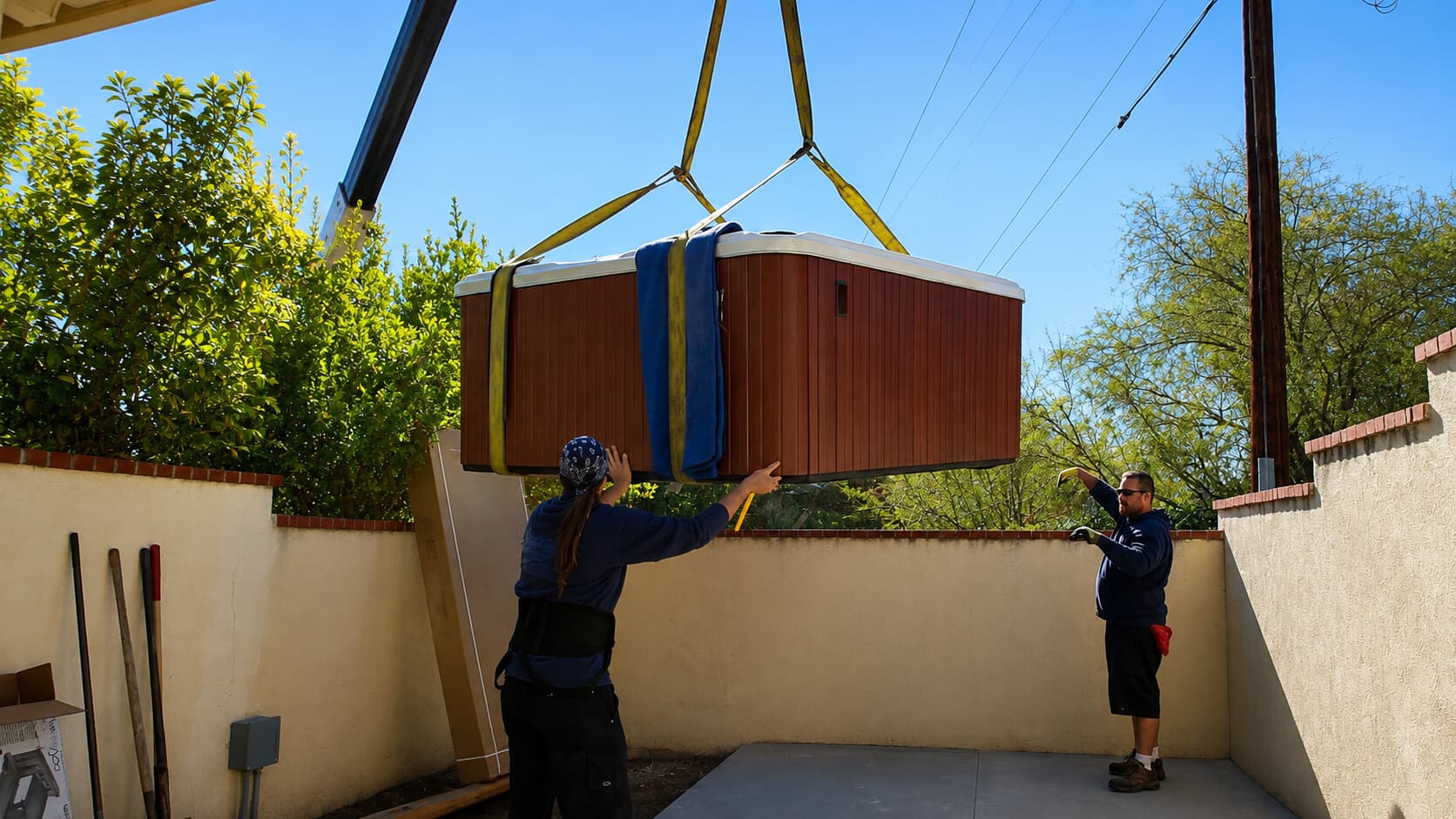 Crane lowering a hot tub into a narrow walled courtyard as two crew members guide it