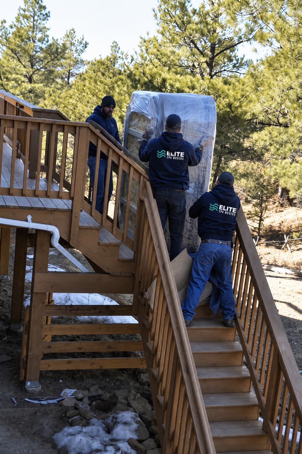 Three-person crew carrying a wrapped hot tub up a wooden deck staircase in snow
